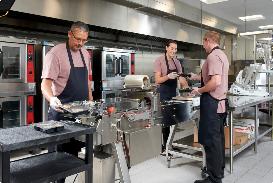 Three kitchen workers using Oliver Speedseal Meal Packaging System MX-2