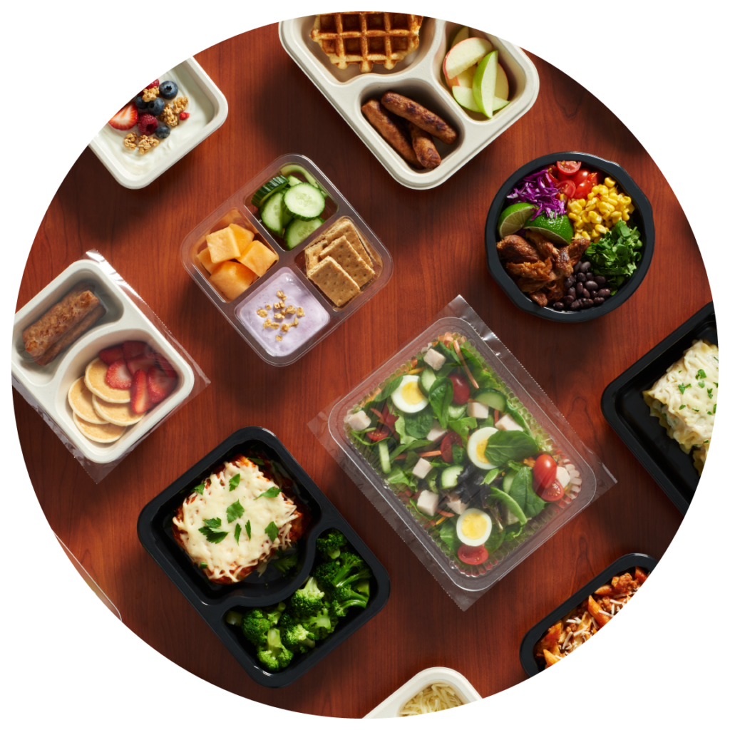 Assortment of plastic and fiber trays, on wooden table, containing various snacks and meals.